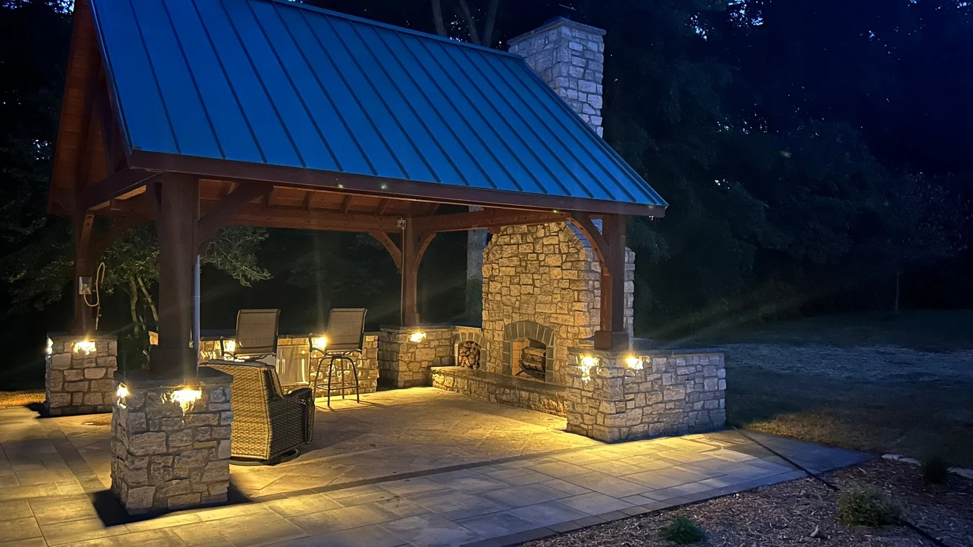 Outdoor pavilion with lighting and stone fireplace in landscaped area at dusk.