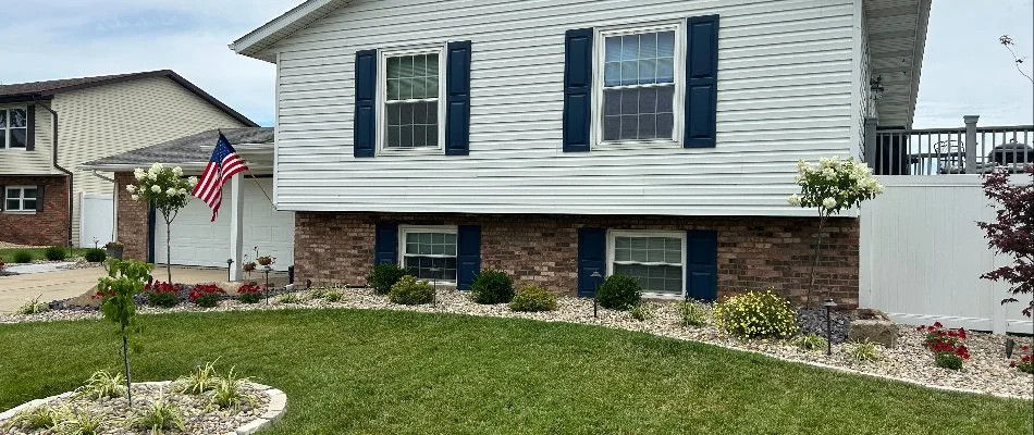 Front view of a split-level home with brick and siding, a landscaped yard with flower beds and young trees, and an American flag on display.